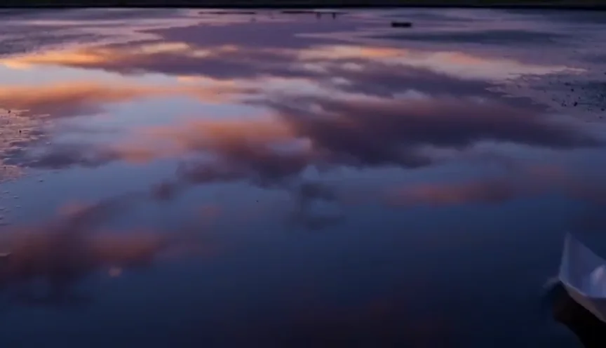 A paper boat drifting across a still puddle at dusk, the sky reflected around it