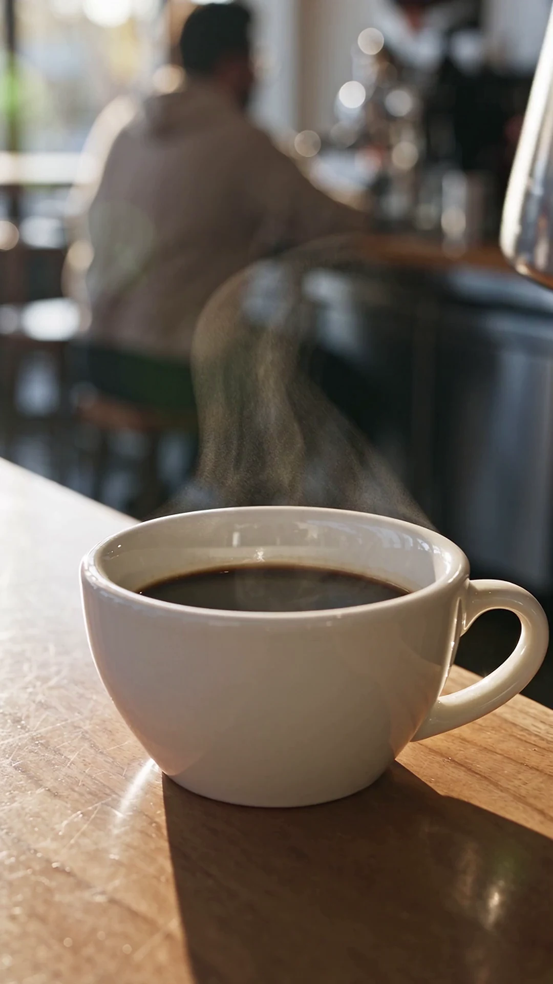 Güneşli bir kafe masasında buharı tüten siyah kahve fincanına çelik bir potdan süt dökülüyor; Premium motorda render edildi